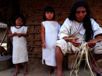 Ein junger Kogi sitzt auf einem Holzschemel und stellt einen Bastkorb her, während Kinder dabei zuschauen